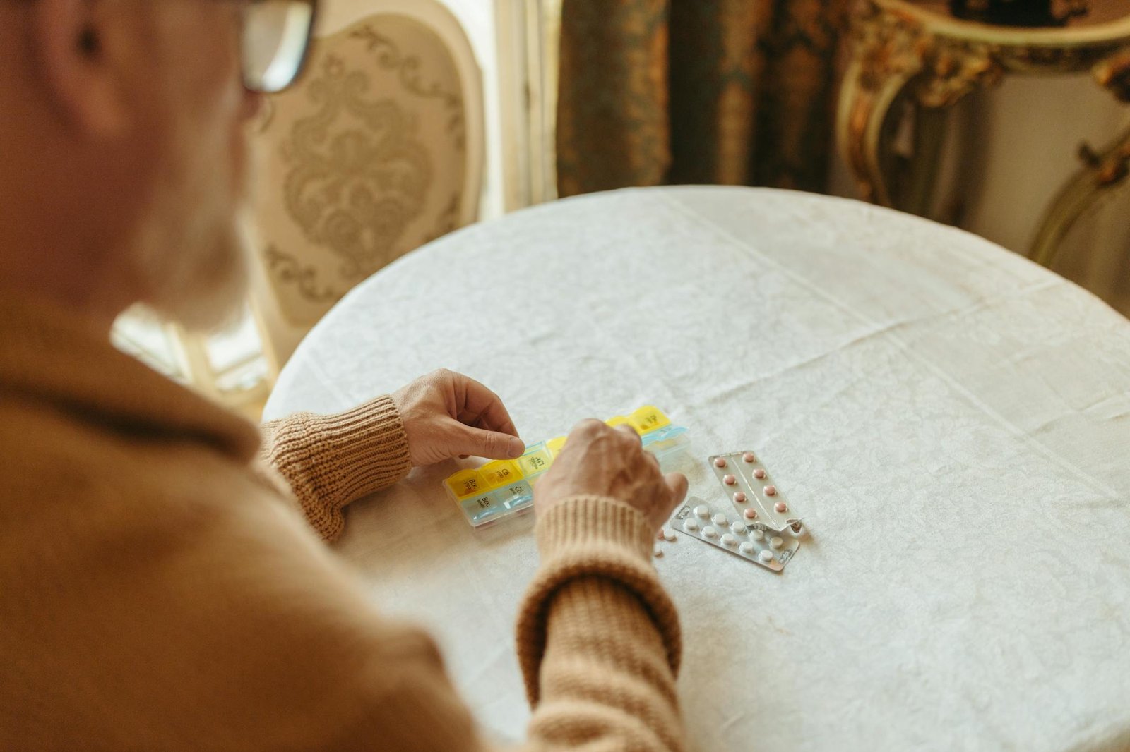 Elderly person sorting daily medication with a pill organizer on a table indoors.